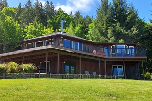 A two-story wooden house with a balcony, surrounded by a well-maintained lawn and dense forest. - Idyllic home with water views, wrap-around deck, hot tub, and firepit - dogs ok (Port Angeles)