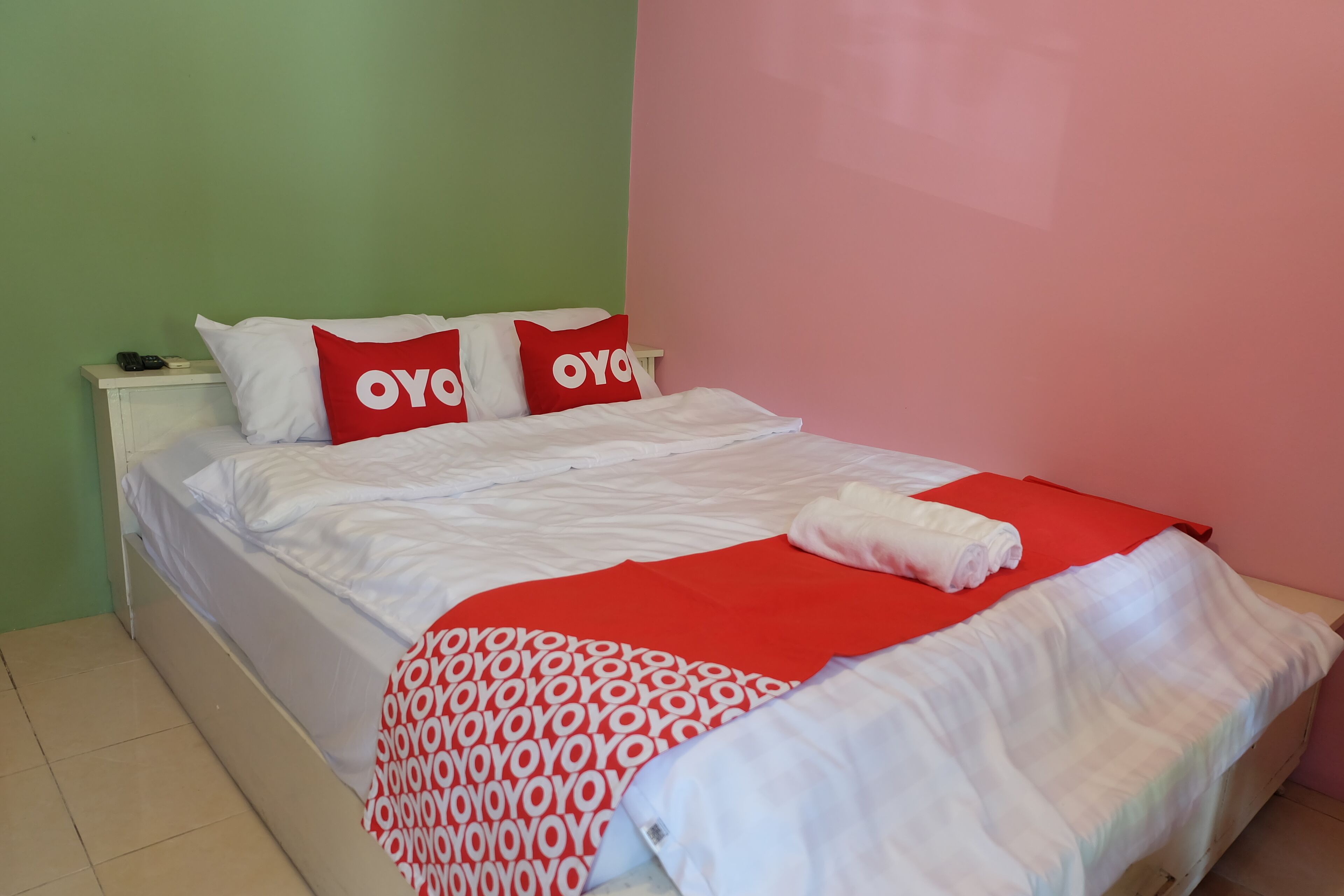 Una cama bien hecha con una colcha de rayas rojas y blancas, dos almohadas con la palabra 'OYO' y una mesita de noche blanca con un teléfono y un control remoto.