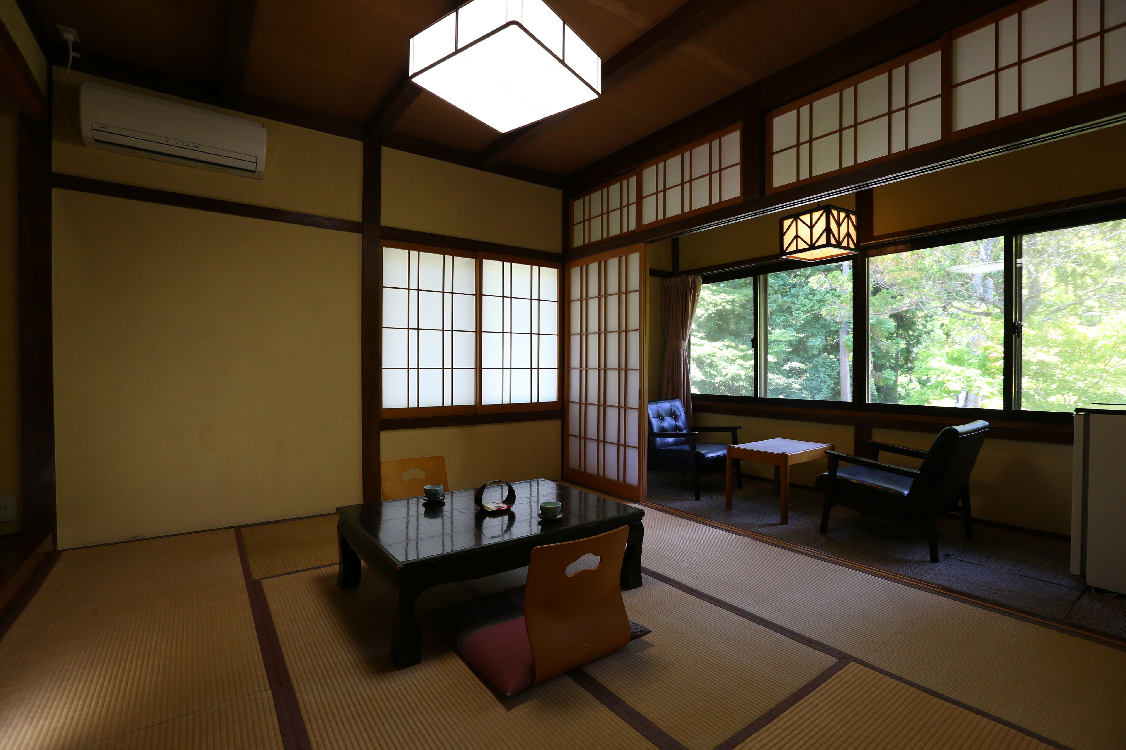 A traditional Japanese room with tatami flooring, a low table with cushions, a sofa, and a view of greenery through large windows.