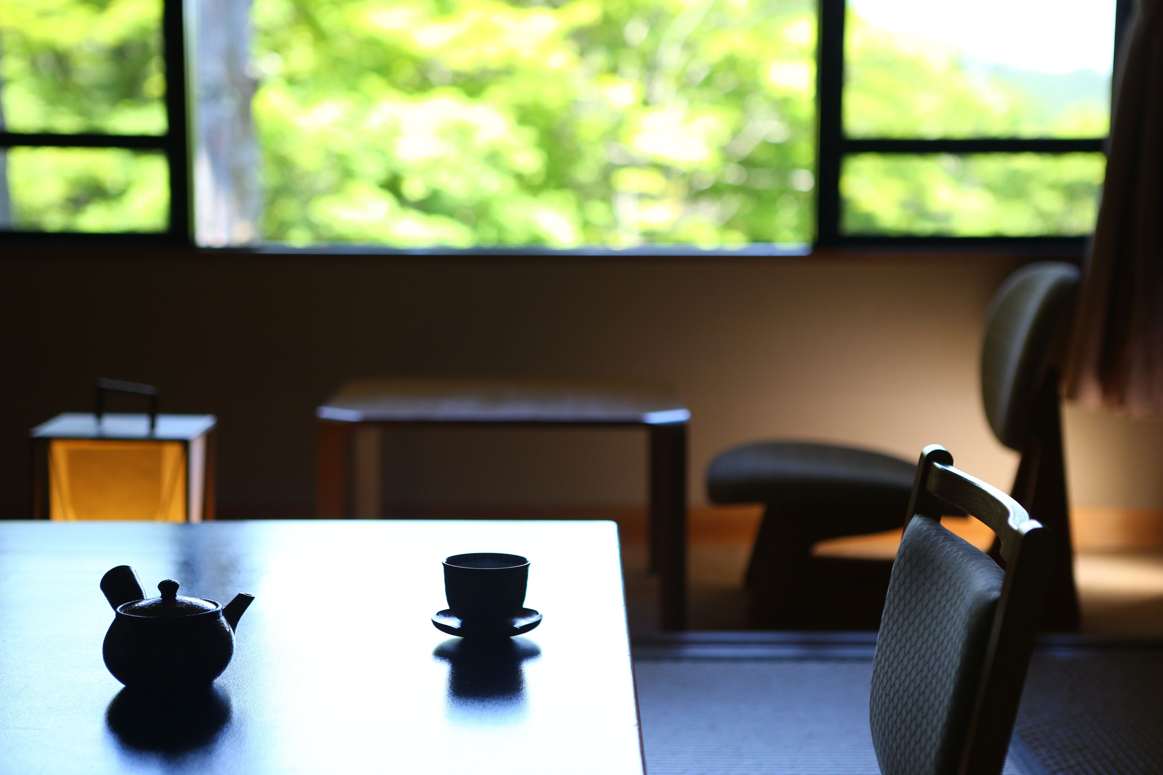 Una mesa con un hervidor y una taza, una silla y un ventanal con vistas al verdor.
