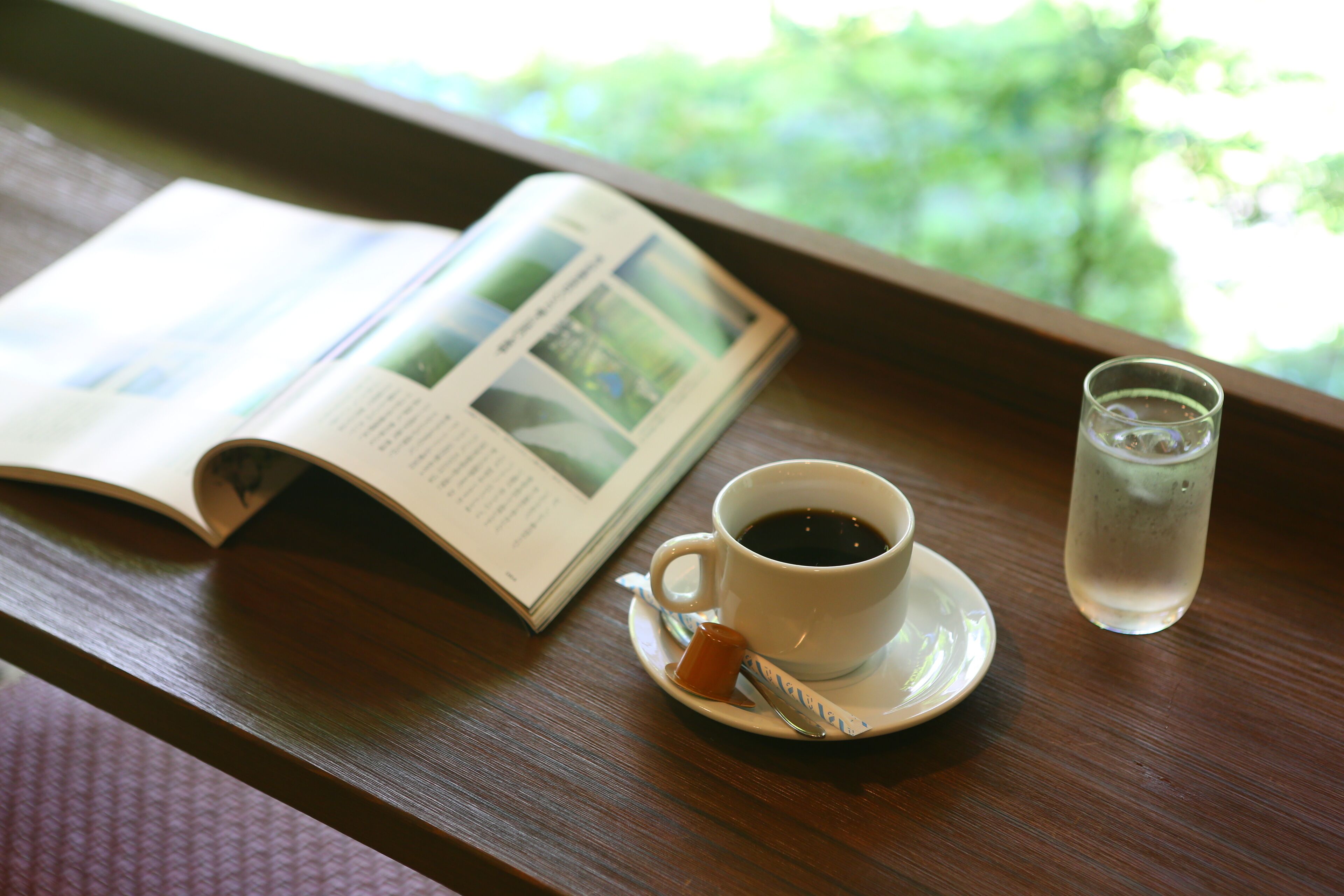 A book, a cup of coffee, and a glass of water on a wooden table by a window.