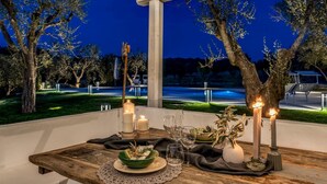 An outdoor dining area with a wooden table set for two, candles, and a view of the surrounding trees and street at night. - Trullo La Sacchina in Ostuni (Ostuni)