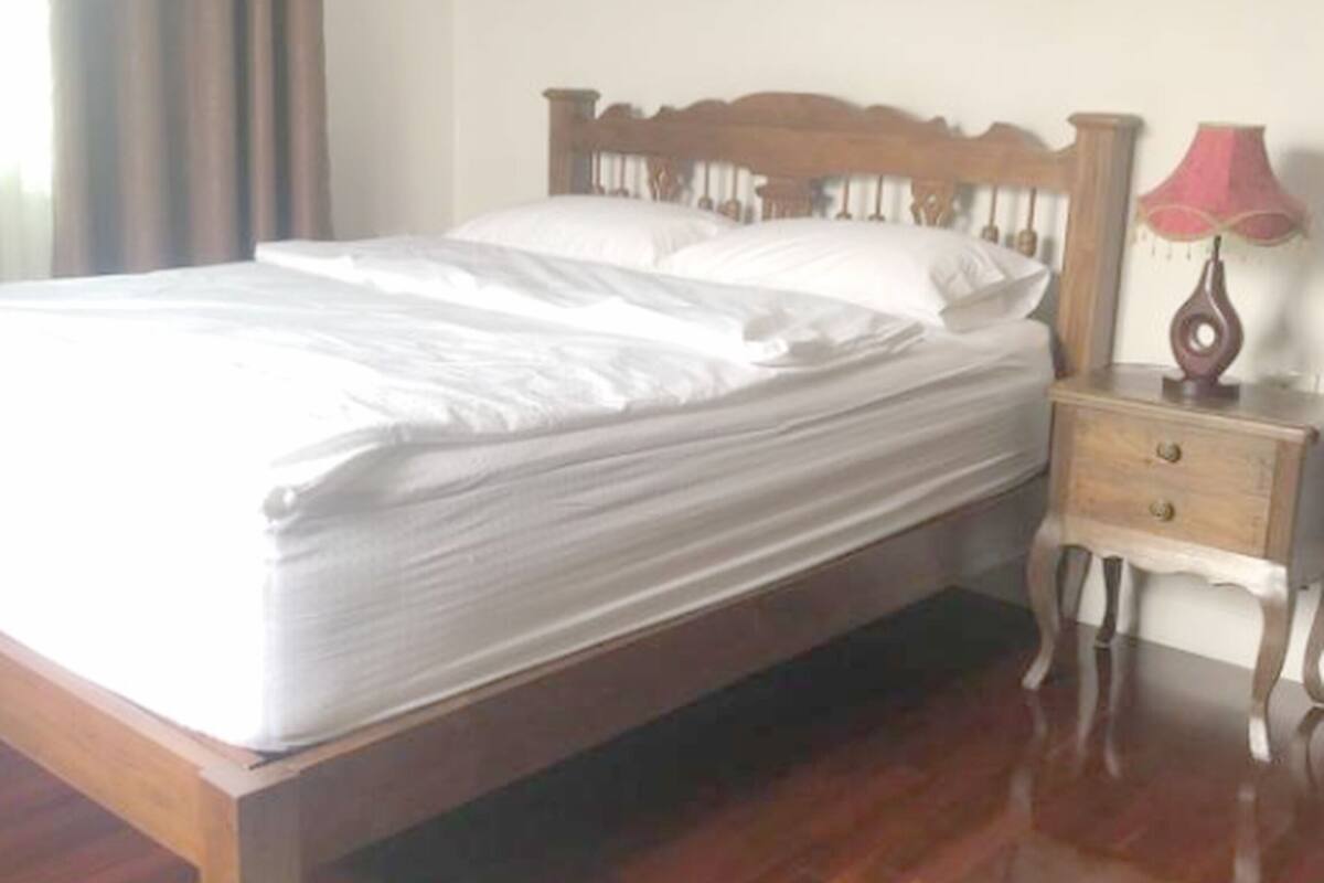 A wooden bed with white bedding in a room with wooden flooring and a nightstand with a lamp.