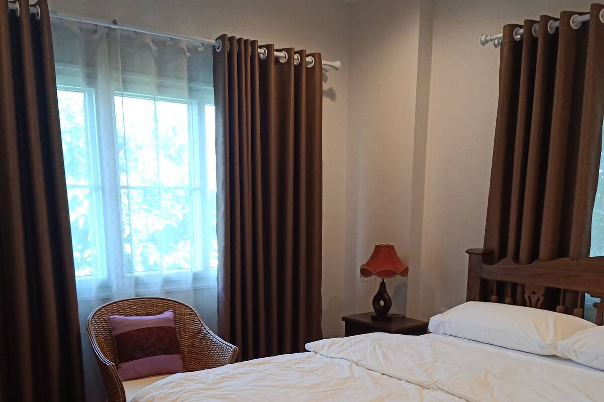 A bedroom with a large bed, a chair, a nightstand, and a window with curtains.