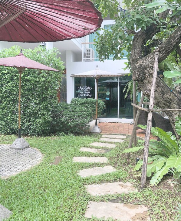 A garden pathway leading to a building with a glass entrance, surrounded by greenery and a red umbrella.