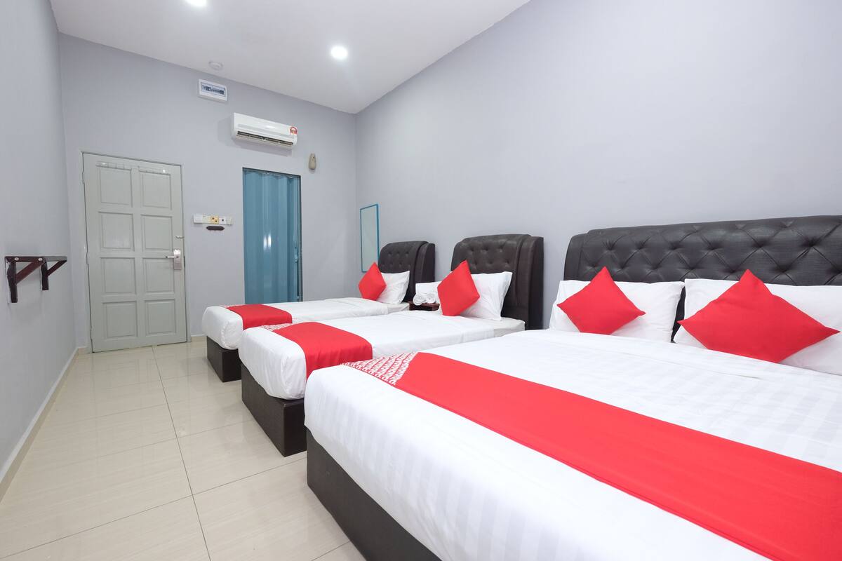A hotel room with three beds, each with white bedding and red accents, a blue door, and an air conditioning unit on the wall.