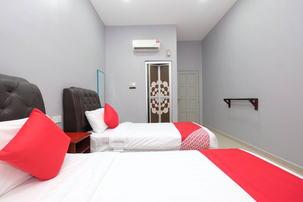 A hotel room with two beds, each with red and white bedding, a grey wall, a white door, and a small wooden shelf.