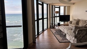 A spacious room with large windows offering a view of the ocean, a modern sofa, a flat-screen TV, and a wooden floor. - Oppulent Apartment on the Beach (Cape Town)