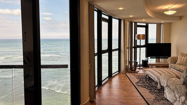 A spacious room with large windows offering a view of the ocean, a flat-screen TV, and a cozy seating area with a patterned rug. - Oppulent Apartment on the Beach (Cape Town)