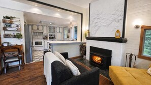 A cozy living room with a fireplace, a sofa, and a dining area with a kitchen in the background. - In the heart of the village, lake, hiking and more (Saint-Adolphe-d'Howard)