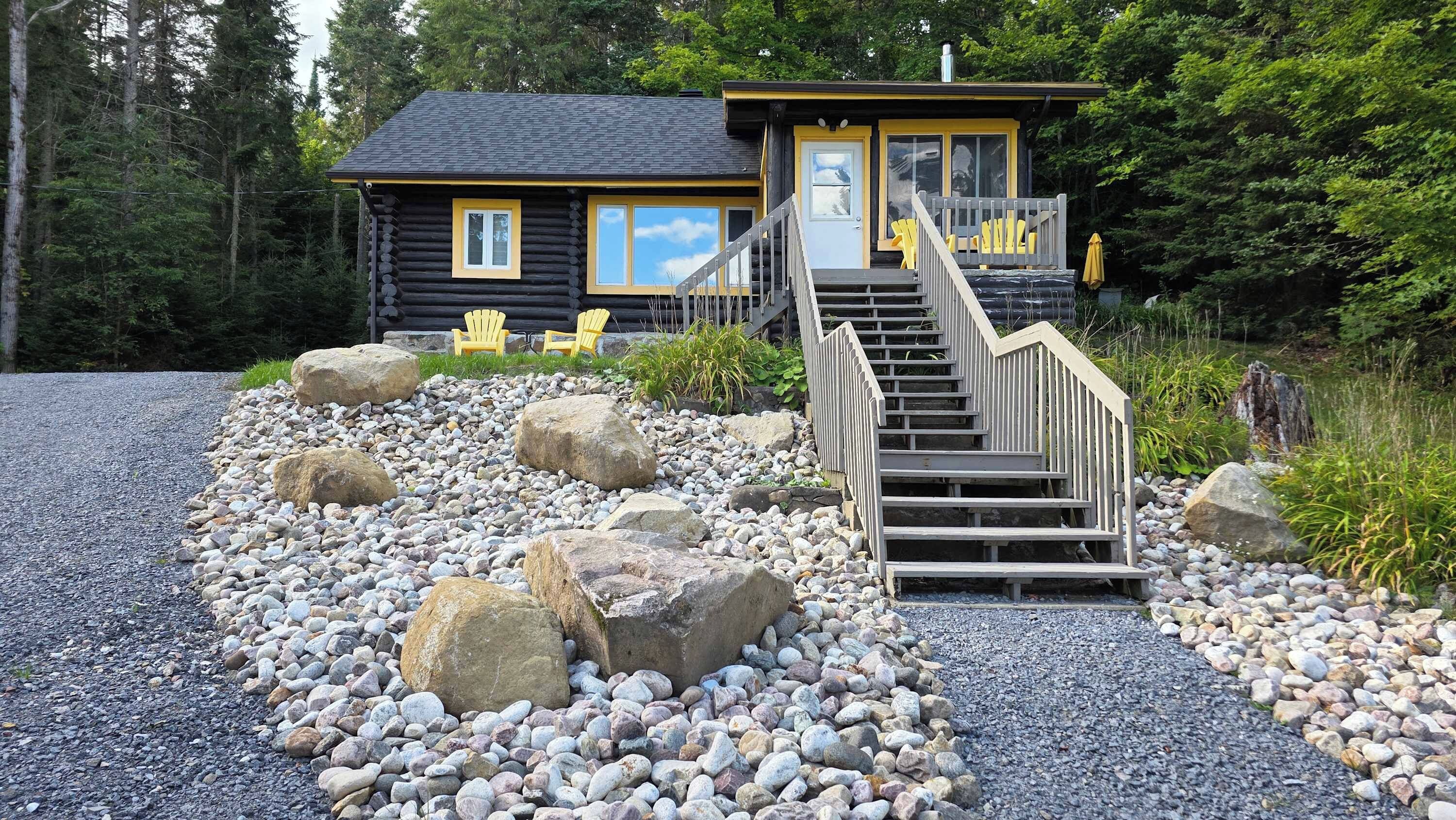 Een chalet met een houten trap die uitkomt op een terras, omgeven door een grindpad met rotsen en begroeiing.