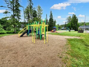 A playground with climbing structures and slides, surrounded by trees and a clear sky. - In the heart of the village, lake, hiking and more (Saint-Adolphe-d'Howard)