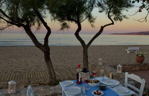 A table set for two with a bottle of red wine, glasses, and plates on a beach with a view of the sea and a sunset.