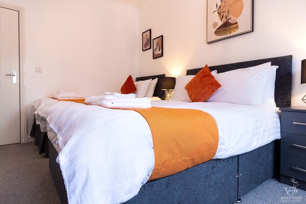 Un lit bien fait, avec une literie blanche et orange, une tête de lit sombre et une table de chevet avec une lampe.