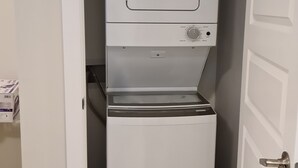 A compact, white laundry machine in a narrow room with a wooden floor. - Creekwood Luxury 1 Bed Lower Level Suite (Edmonton)