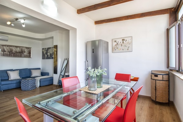A modern living room with a glass dining table, red chairs, a blue sofa, and a large stainless steel refrigerator.