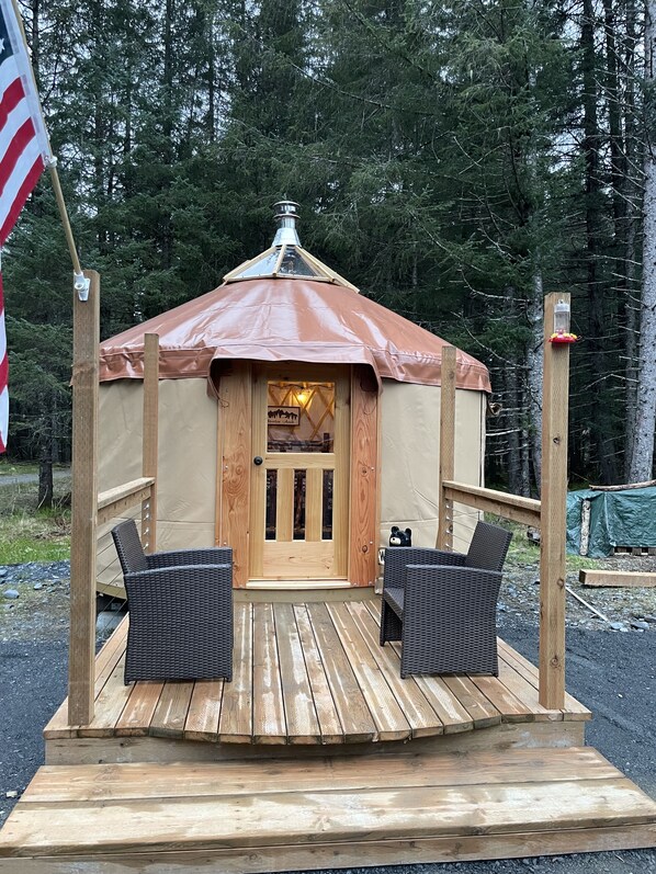 Une yourte en bois avec un toit brun, une porte portant une inscription et deux chaises en osier sur une terrasse en bois.