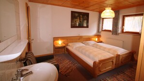 Un dormitorio con cabecera de madera, dos camas, una mesita de noche con lámpara, una ventana con cortinas, un cuadro en la pared y un baño con lavamanos y bañera.
