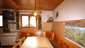 Una mesa de comedor de madera con bancos, un televisor sobre un mueble bajo, una ventana con cortinas y una pared con una cruz y un cartel.