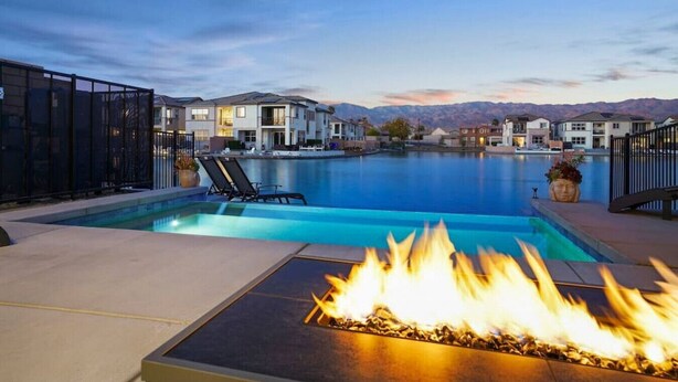 A swimming pool with lounge chairs and a fire feature, surrounded by modern houses and mountains in the background.
