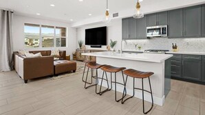 A modern kitchen with grey cabinets, a large island, and a flat-screen TV. There is a seating area with a sofa and a coffee table, and a large window offering a view of the outside.