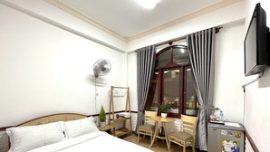 Une chambre d’hôtel équipée d’un lit, d’un bureau, d’une chaise, d’une petite table, d’un ventilateur, d’un réfrigérateur et d’une fenêtre avec des rideaux.