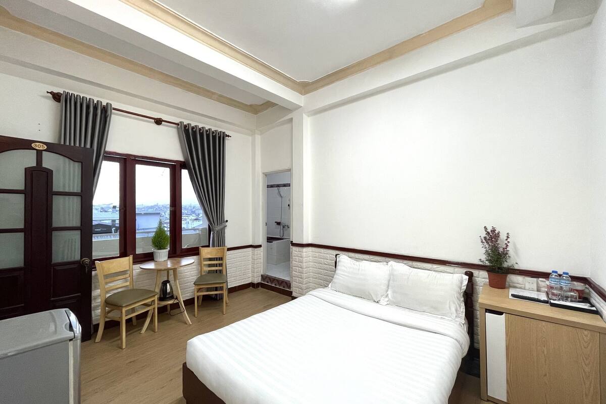 A hotel room with a bed, a desk, a chair, a potted plant, and a view of the city.