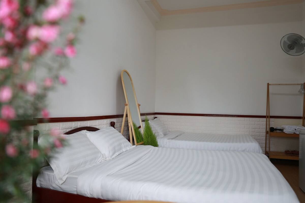 A hotel room with two beds, a wooden headboard, a small mirror, a fan, and a shelf with folded towels.