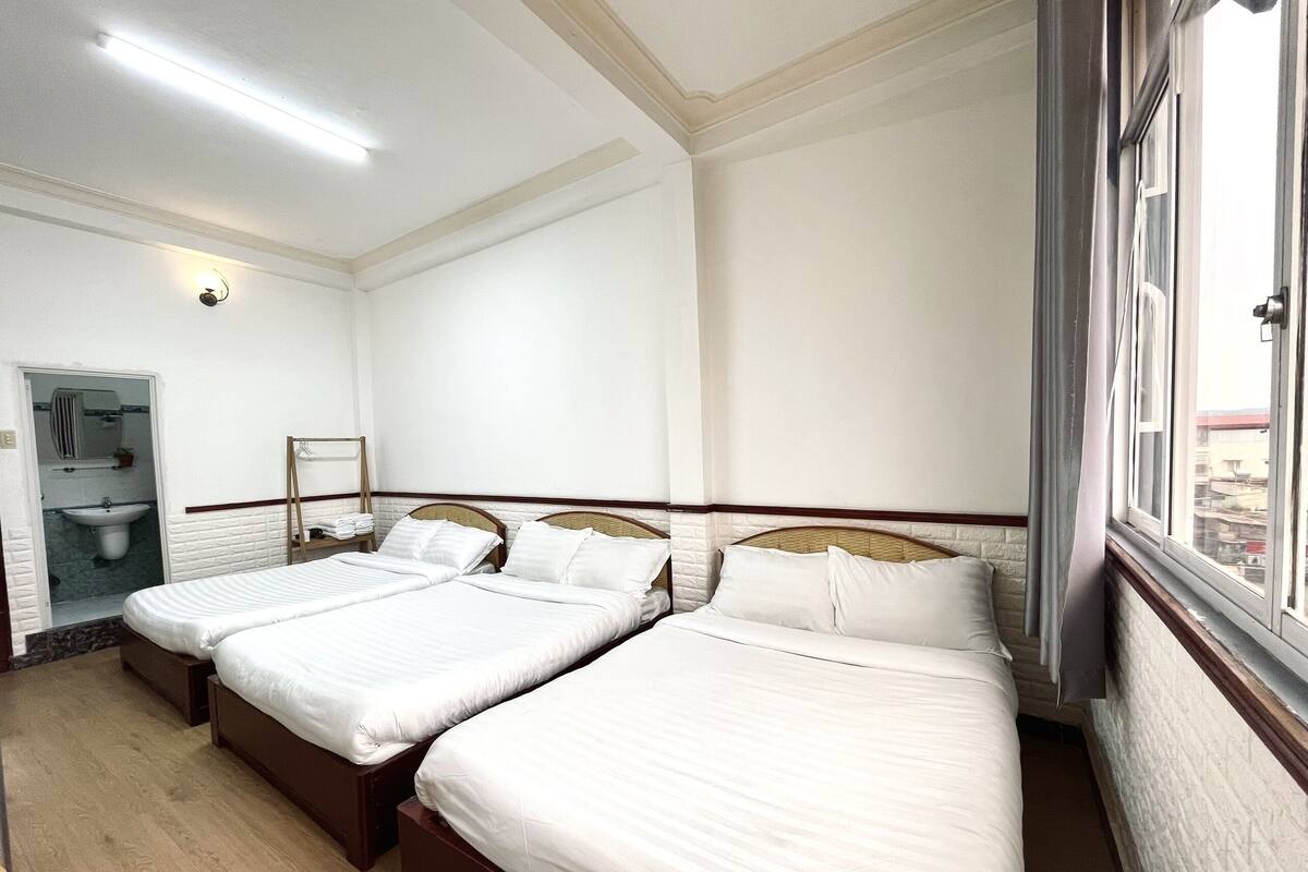 A hotel room with three single beds, a bathroom with a sink and mirror, and a window with a view of buildings.