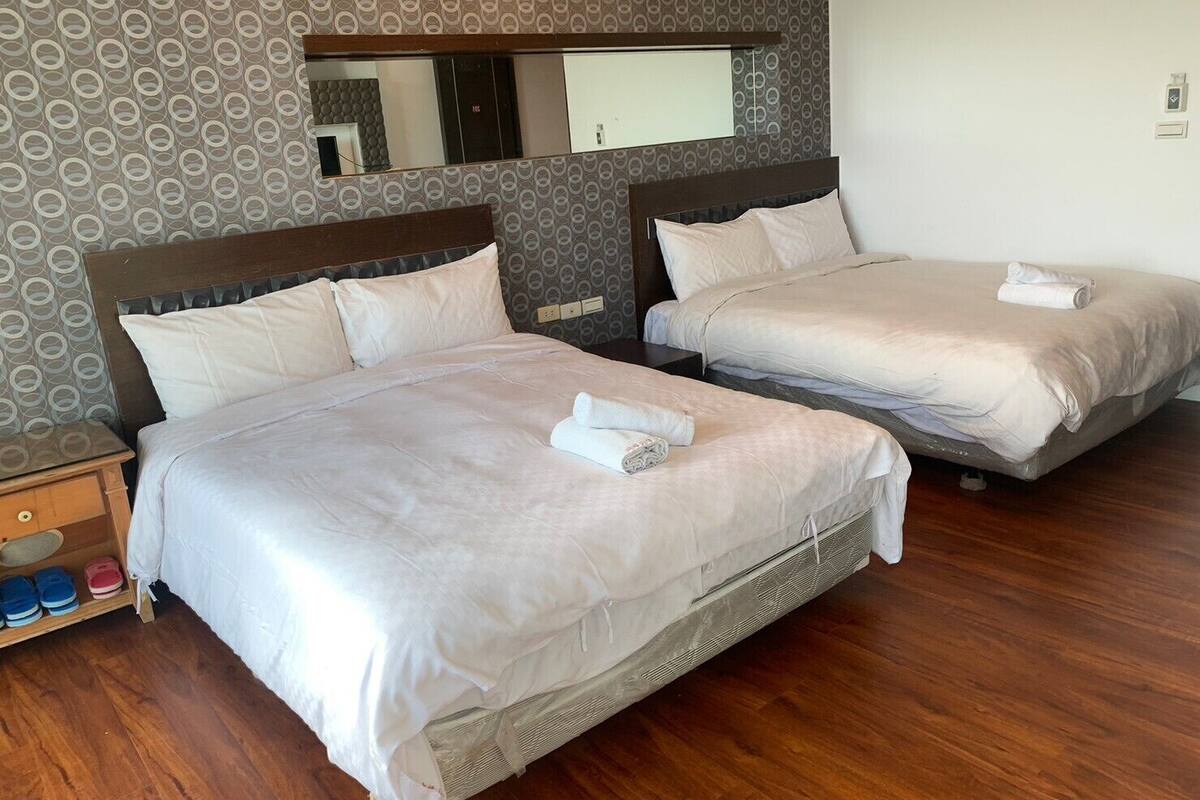 A hotel room with two beds, a wooden nightstand with slippers, and a mirror above the beds.