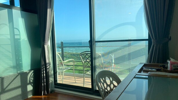 A room with a large window offering a view of the ocean and a balcony with chairs. - Happiness Inn (Hengchun)