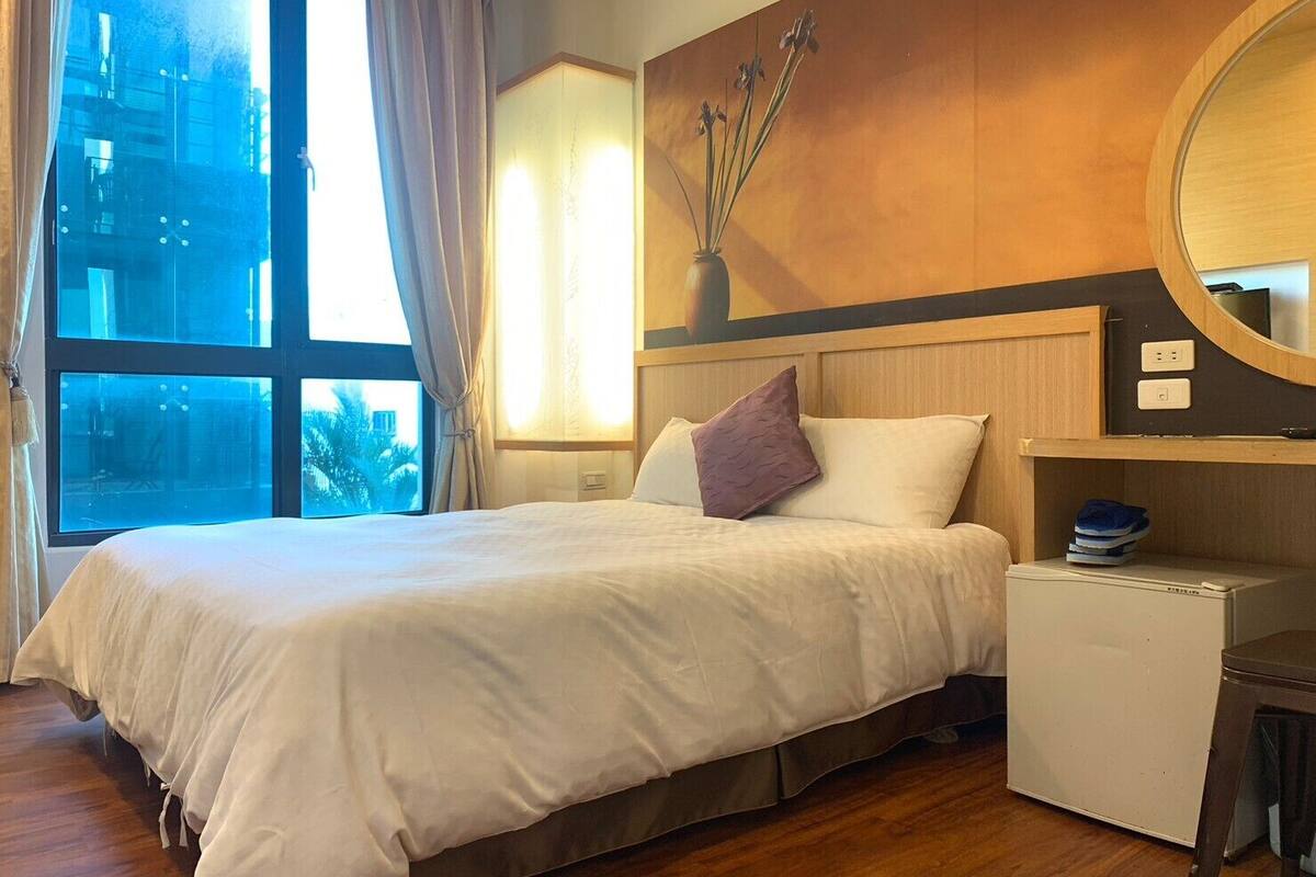 A hotel room with a large bed, a small desk, a mirror, and a window with curtains.