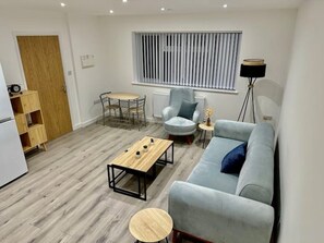 Una sala de estar moderna con un sofá gris, una mesa de centro de madera, un sillón gris y una pequeña mesa redonda. Hay una ventana con persianas, un radiador y una puerta de madera.