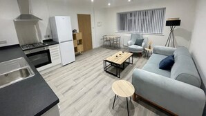 A modern kitchen with a stove, oven, and white refrigerator. There is a dining area with a table and chairs, a light blue sofa, and a small round table. - A newly built 2023 apartment (Oxford)