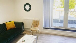 Un salon avec un canapé vert foncé, un coussin jaune, une chaise en bois et une table basse blanche. Une grande fenêtre, ornée de rideaux, offre une vue sur l’extérieur.