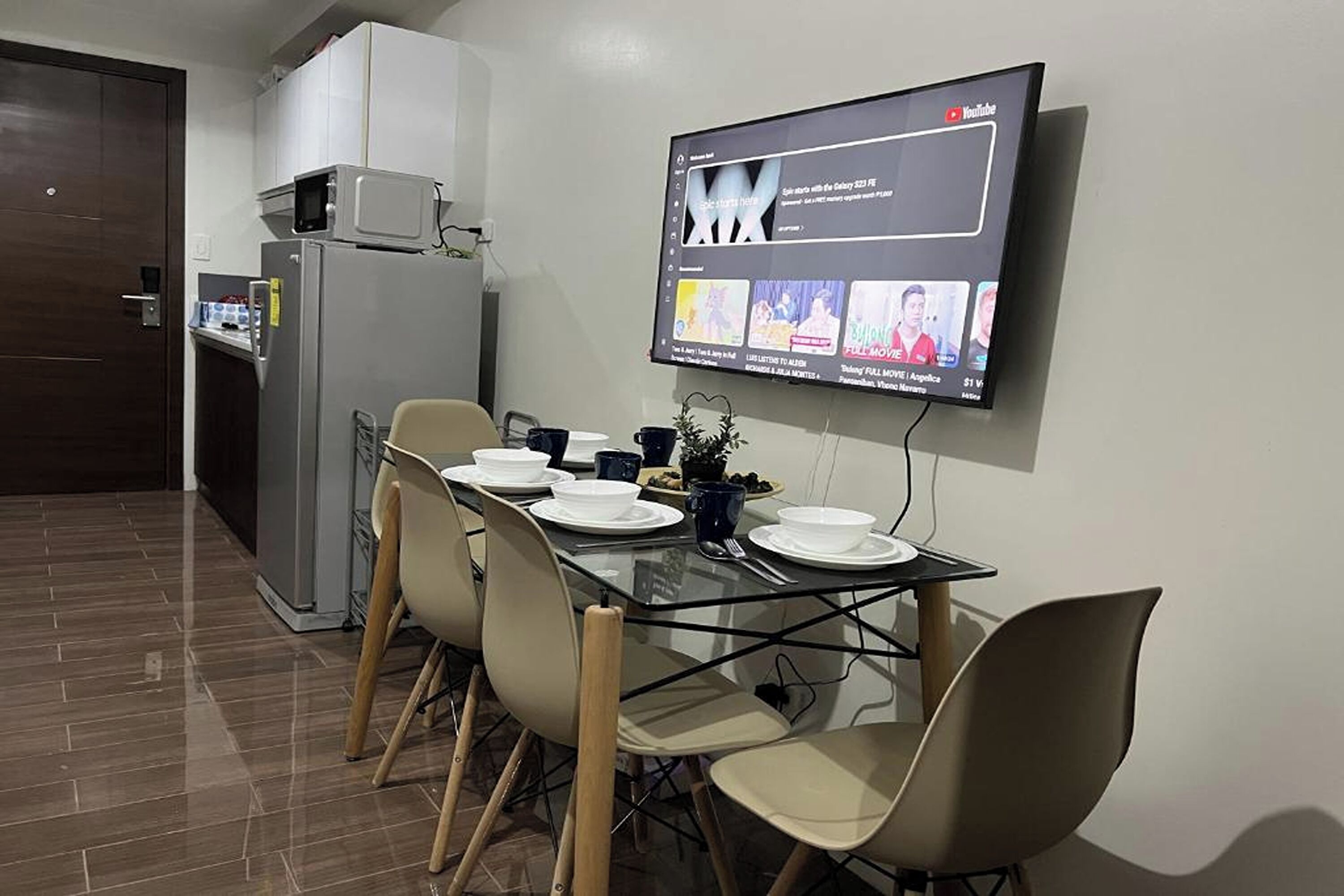 A modern kitchen with a dining area. There is a flat-screen TV mounted on the wall displaying YouTube content. The kitchen features a refrigerator, microwave, and a small cabinet. The dining table is set with plates, bowls, and cups.