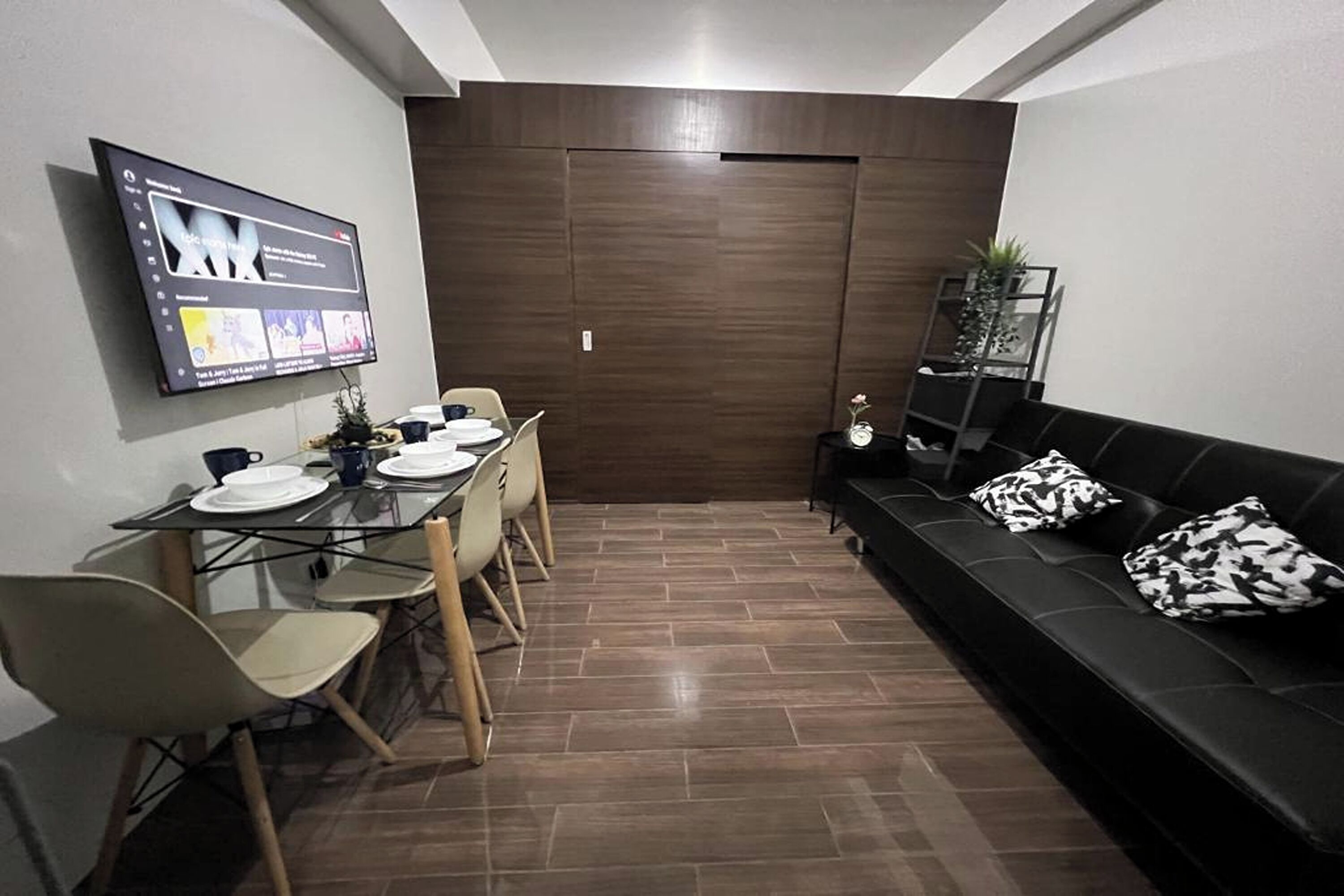 A modern living room with a black leather sofa, a dining table with chairs, and a flat-screen TV mounted on the wall.