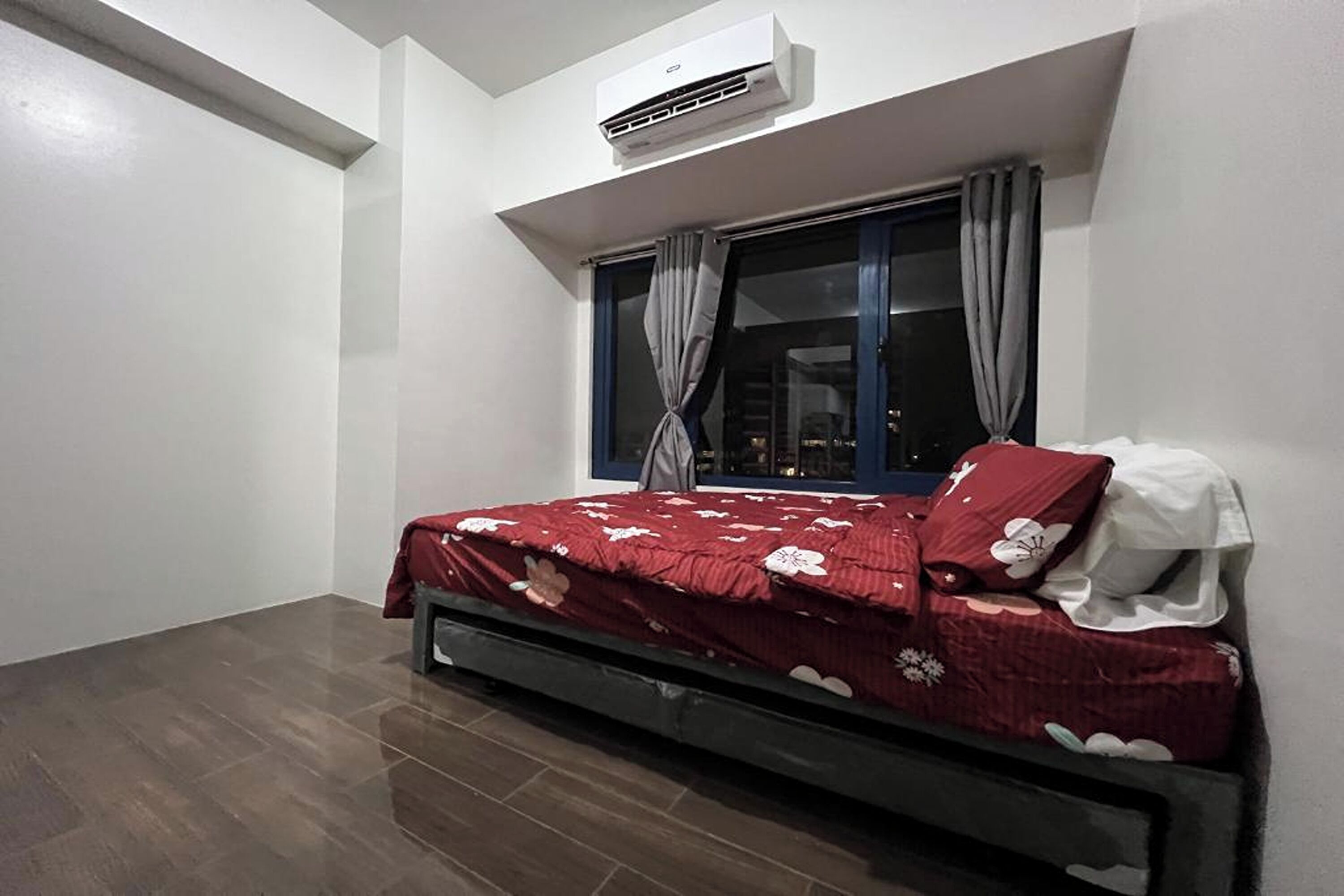 Een slaapkamer met een bed, een airconditioner, een raam met gordijnen en uitzicht op de stad 's nachts.