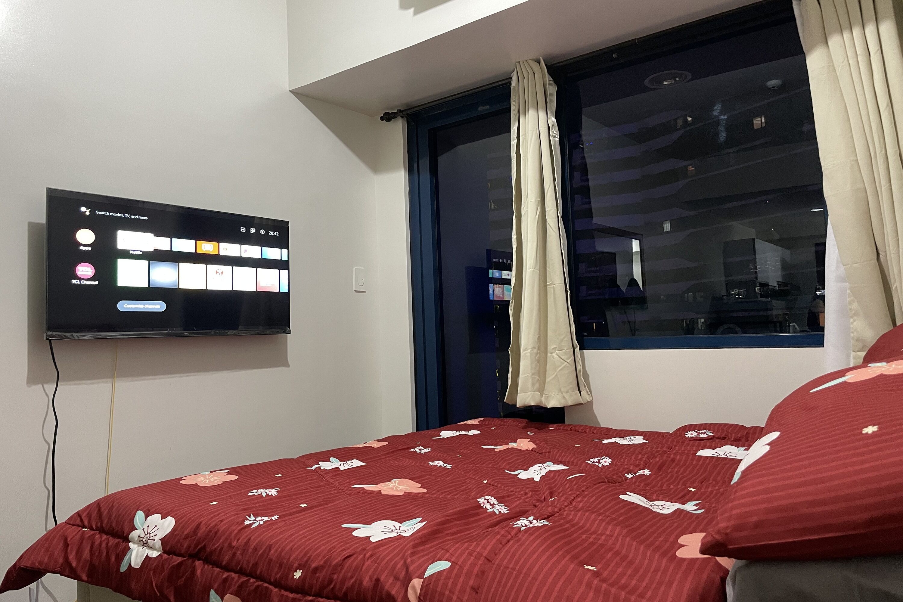 Une chambre avec un téléviseur fixé au mur, une fenêtre avec des rideaux et un lit avec une literie à motifs floraux.