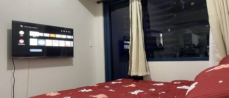 Une chambre avec un téléviseur fixé au mur, une fenêtre avec des rideaux et un lit avec une literie à motifs floraux.