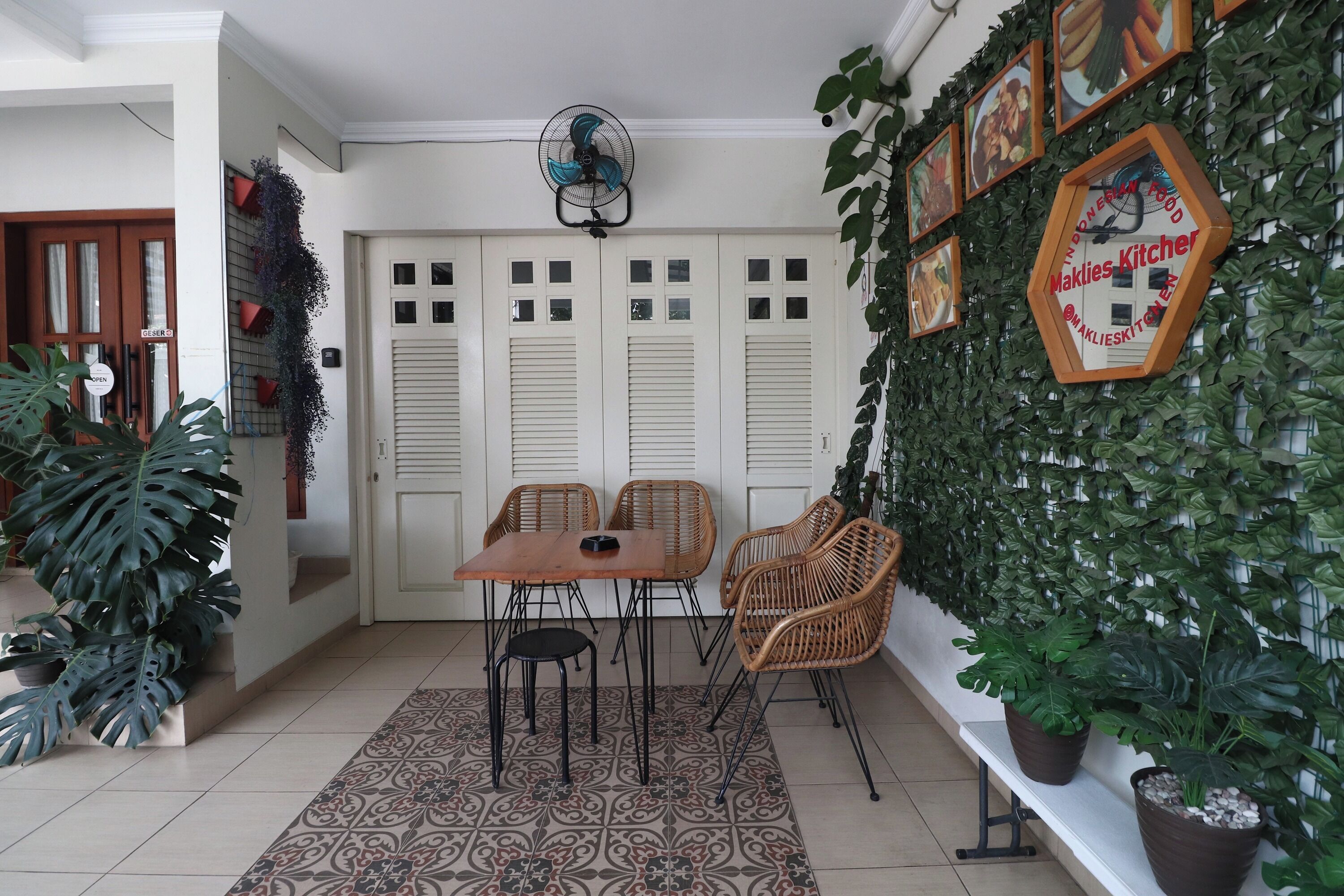 En spiseplads med bord og stole, en ventilator samt en væg pyntet med planter og billeder i rammer.