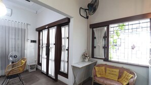 A room with a sofa, a chair, a small table with a vase, a fan, and a window with curtains. - Bisma Balcony Homestay (Pogung Lor)
