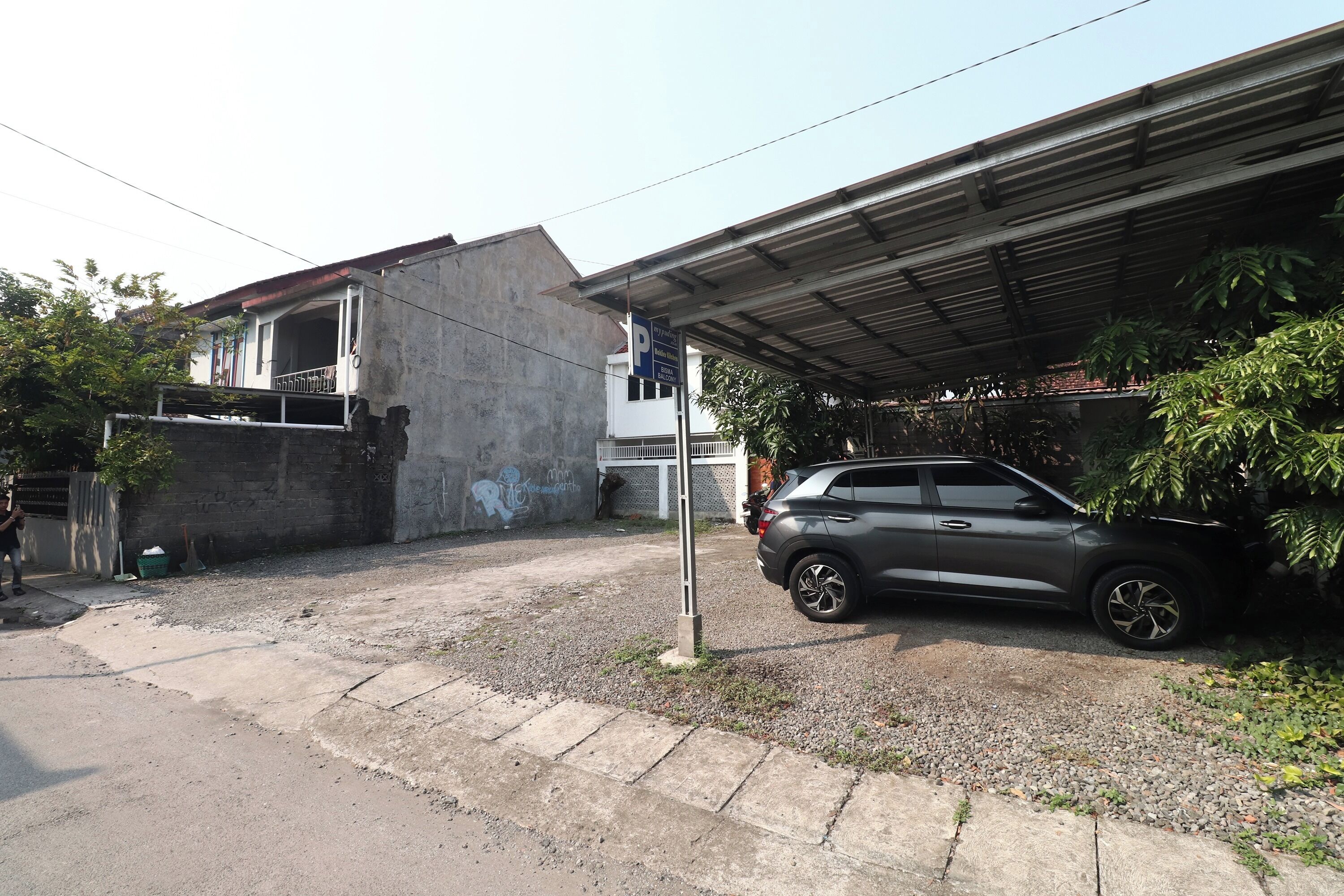 Zona residencial amb un cotxe aparcat, un edifici de formigó i una estructura coberta.