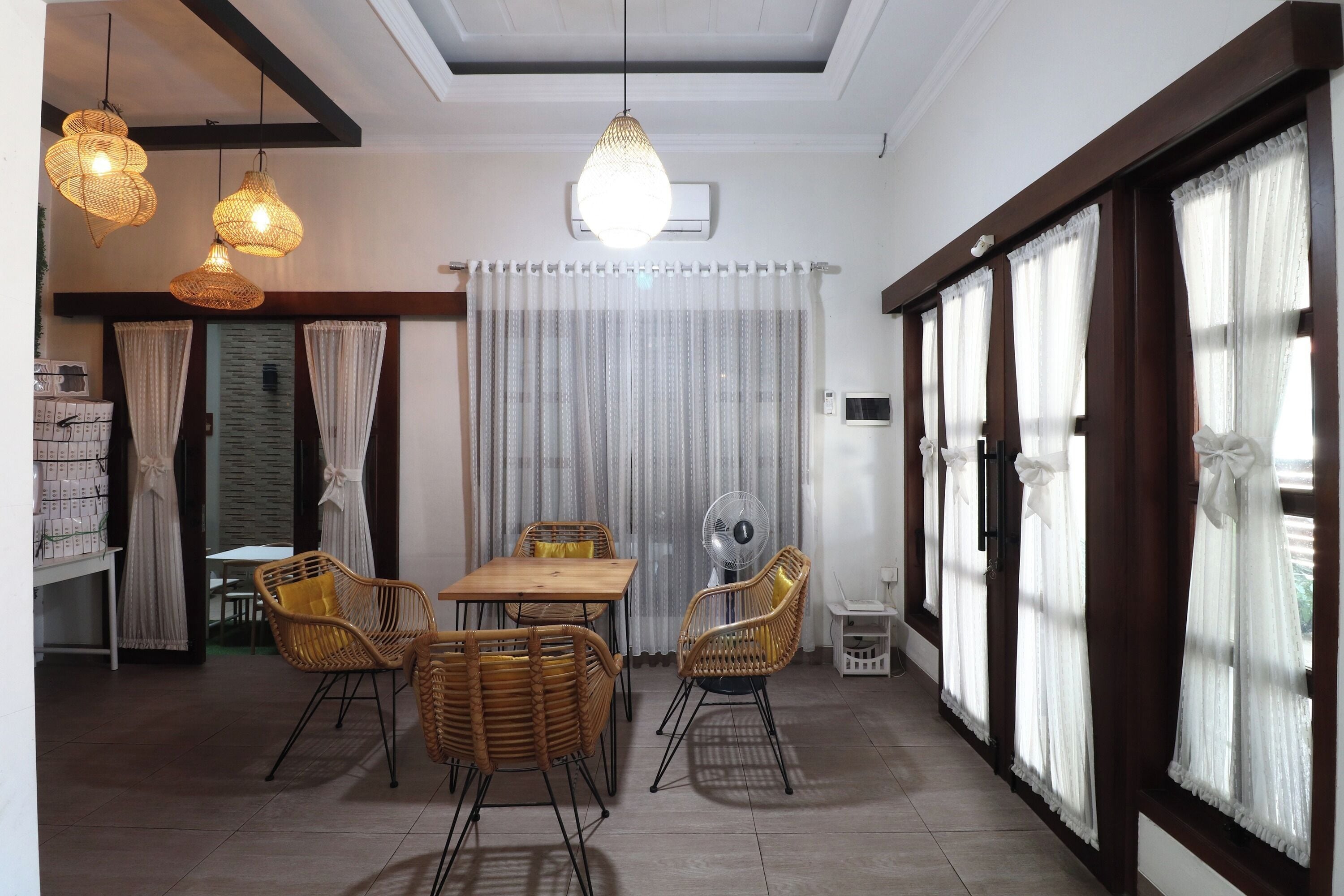 Un comedor con mesa de madera y sillas de mimbre, ventilador de techo y ventana con cortinas.