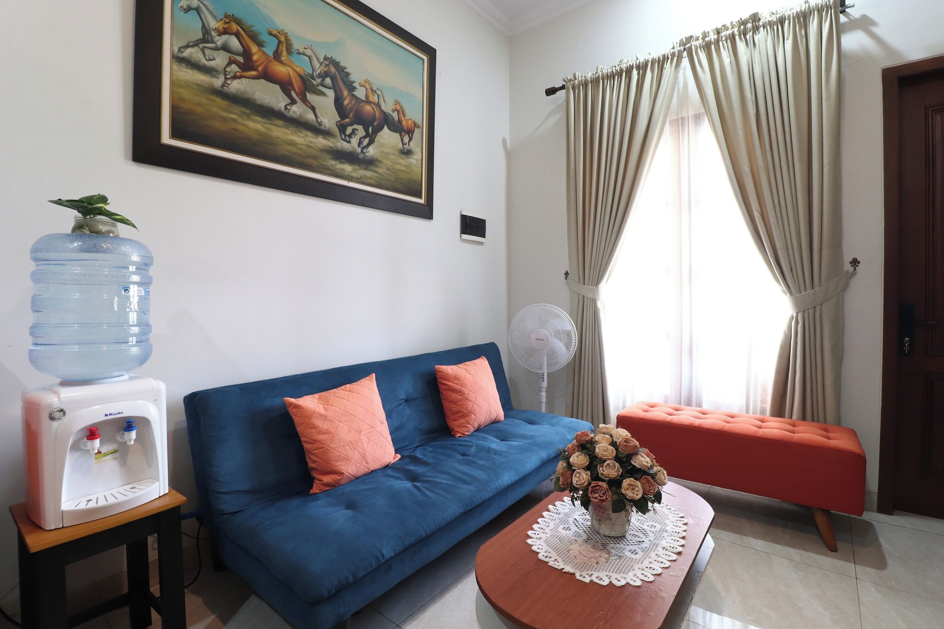 A living room with a blue sofa, a water dispenser, a fan, and a painting of horses.