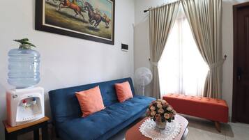 A living room with a blue sofa, a water dispenser, a fan, and a painting of horses.