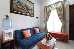 Un salon avec un canapé bleu, un distributeur d’eau, un ventilateur et un tableau représentant des chevaux.