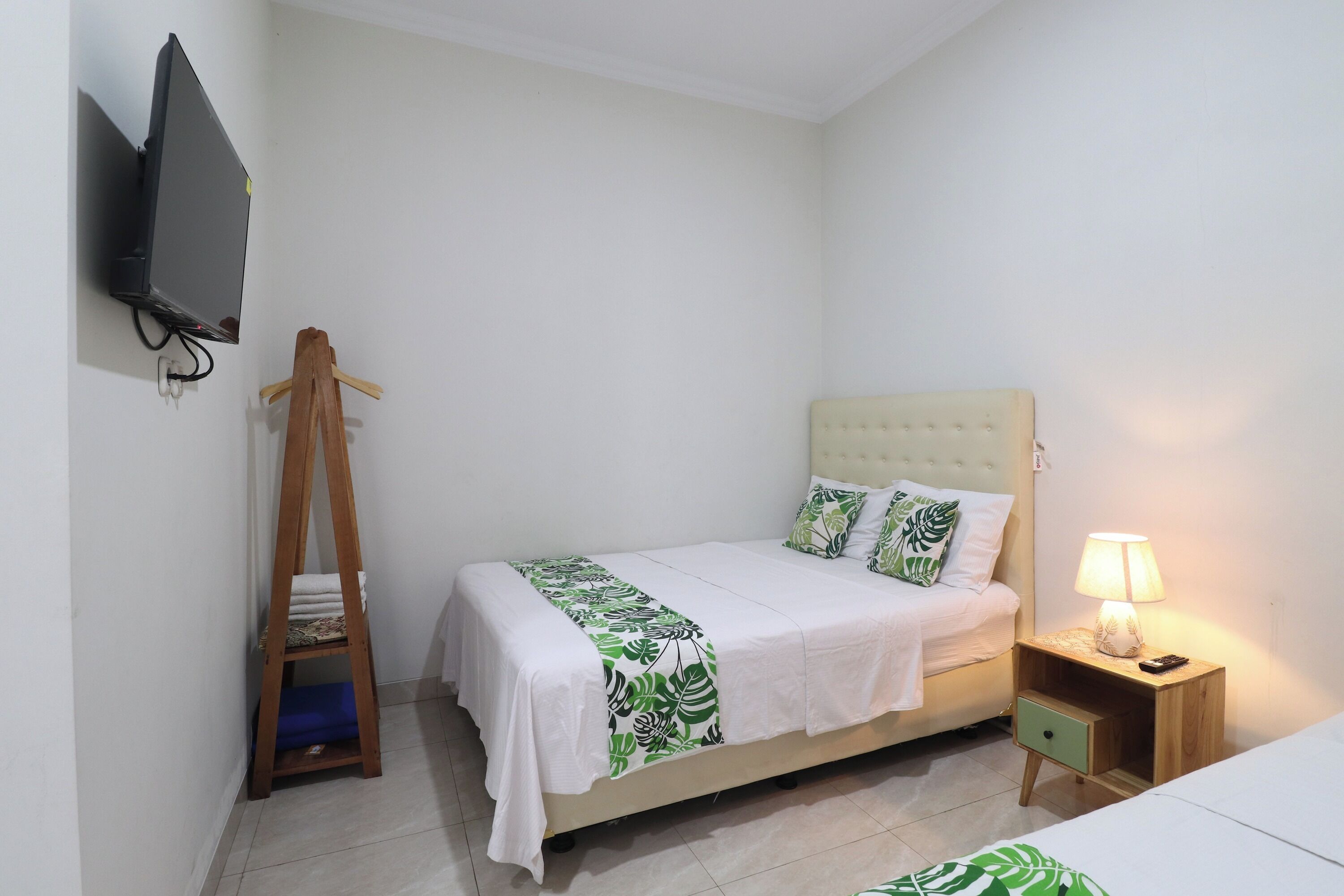 Un lit simple avec une couvre-lit blanche et verte à motifs de feuilles, un cintre en bois, un téléviseur fixé au mur et une table de chevet avec une lampe.