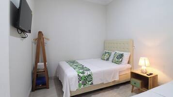 Un lit simple avec une couvre-lit blanche et verte à motifs de feuilles, un cintre en bois, un téléviseur fixé au mur et une table de chevet avec une lampe.