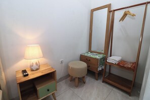 A compact room with a wooden nightstand, a lamp, a small stool, a mirror, and a wooden wardrobe with a hanging rack. - Bisma Balcony Homestay (Pogung Lor)